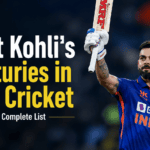 Virat Kohli wearing his blue India jersey celebrating on the field with a teammate, looking up with his helmet removed and bat raised.